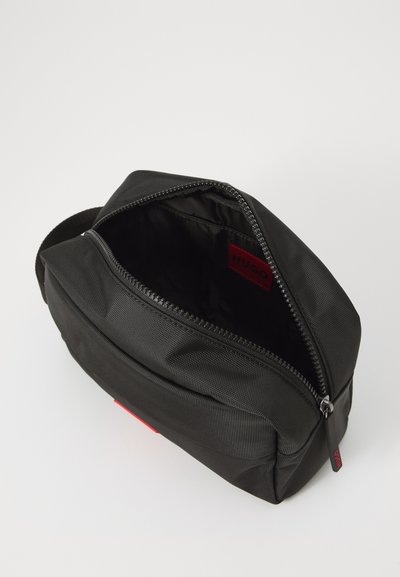 Trousse de toilette en nylon noir avec fermeture éclair, dotée d'une étiquette intérieure rouge et d'une surface extérieure texturée.