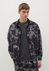 Giacca a track nera e grigia a fantasia con zip, polsini e vita elastici, indossata sopra una maglietta grigia scura. Presenta un logo sul lato sinistro del petto.