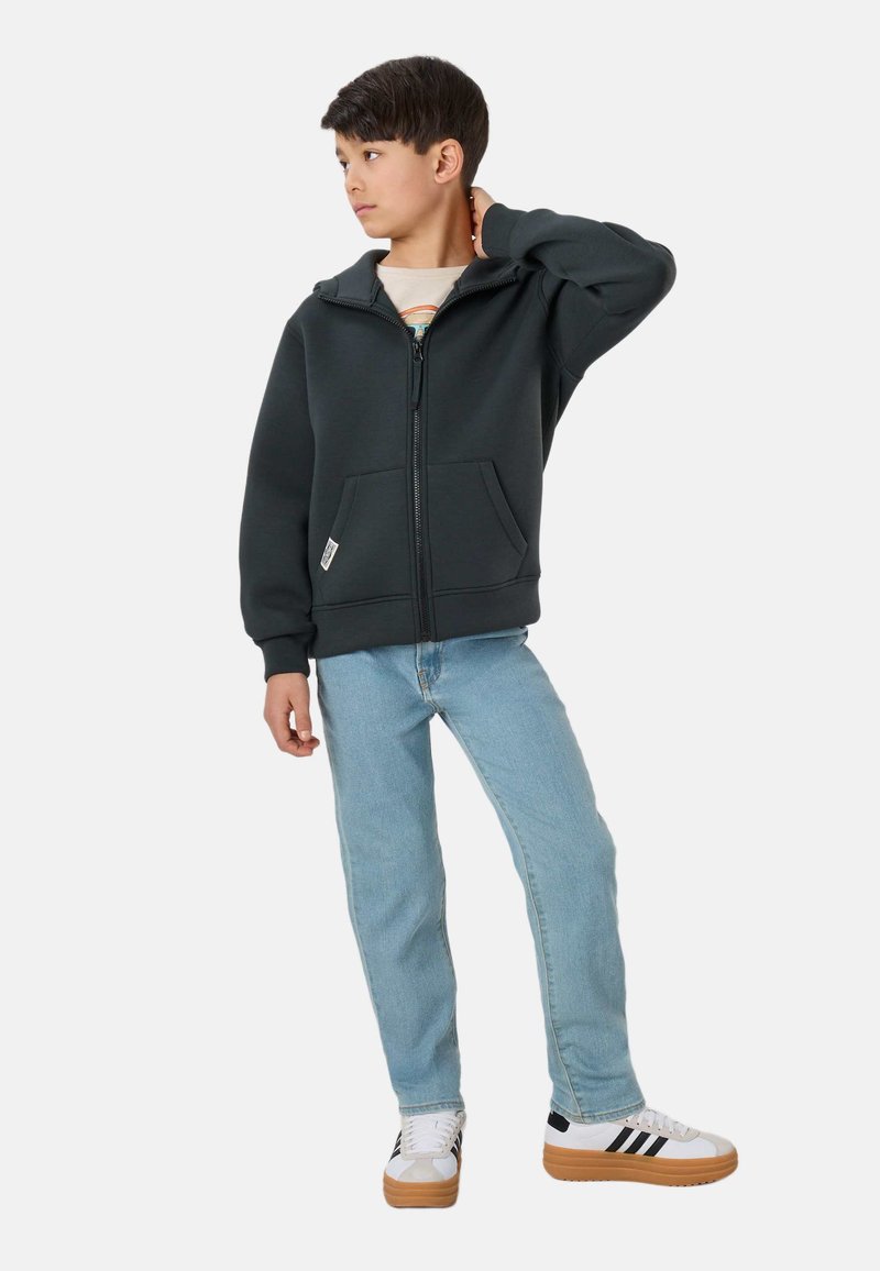 Garçon debout, main sur la nuque, portant un sweat à capuche zippé foncé, un jean bleu clair et des baskets blanches avec des rayures noires.