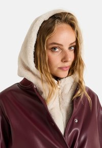 Chaqueta de imitación de cuero burdeos con capucha forrada de piel sintética blanca, cremallera plateada y botones a presión; corte ligeramente holgado, textura suave.