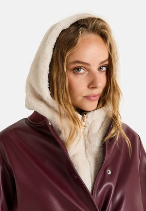 Chaqueta de imitación de cuero burdeos con capucha forrada de piel sintética blanca, cremallera plateada y botones a presión; corte ligeramente holgado, textura suave.
