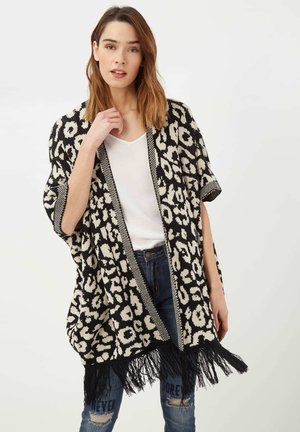 Poncho estilo kimono con patrón en blanco y negro, con dobladillo con flecos, mangas sueltas y corte recto. Llevado sobre una blusa blanca y jeans.