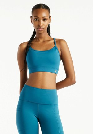 Kvinde med flettet hår iført en blå sports-bh og højtaljede leggings står foran en ensfarvet hvid baggrund.