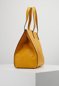 Gul læder tote taske med en struktureret overflade, triangulær form og sorte detaljer. Har to lange, robuste håndtag.