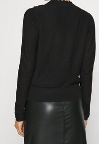 Personne portant un pull noir à manches longues avec col festonné et une jupe en cuir noir, vue de dos sur un fond uni.