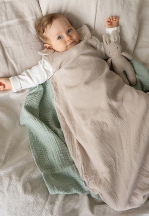 Baby liegt auf dem Bett, eingewickelt in einen beigen Schlafsack mit weißem Langarmshirt, neben gestricktem grauem Hasenspielzeug auf einer grünen Decke.