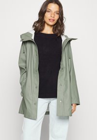Πράσινο αδιάβροχο αδιάβροχο με κουμπιά σνάπ, ρυθμιζόμενη κουκούλα και άνετη εφαρμογή. Συνδυάζεται με μαύρο πουλόβ�ερ και ανοιχτό μπλε τζιν.