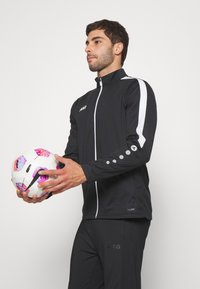 Schwarze Sportjacke mit weißen Akzenten, Reißverschlussfront und gepunkteten Details an den Ärmeln. Das Modell hält einen bunten Fußball.