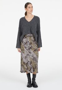 Grauer, weicher, übergroßer Pullover mit V-Ausschnitt und langen Ärmeln, kombiniert mit einem gemusterten, bodenlangen Rock in Erdtönen und schwarzen Knöchelstiefeln.