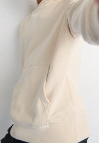 Beige hoodie made of soft fabric, featuring a kangaroo pocket, long sleeves, and a small logo on the chest. Smooth texture and relaxed fit.