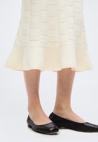 Jambes portant une jupe texturée crème longueur genou associée à des chaussures plates noires simples sur fond blanc.