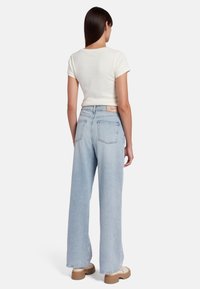 Jean large bleu clair en denim, avec une taille haute, deux poches arrière et une étiquette beige sur la ceinture. Accompagné d'un haut blanc.