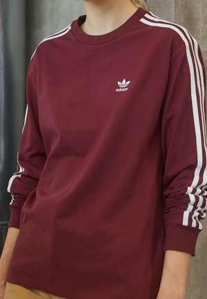 Personne portant un sweat-shirt Adidas bordeaux à manches longues avec des bandes blanches sur les manches et un logo sur la poitrine, debout devant un fond neutre.