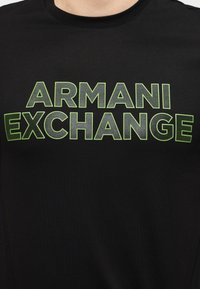 Μαύρο μπλουζάκι με στρογγυλή λαιμόκοψη και έντονο λογότυπο "ARMANI EXCHANGE" σε μεγάλα γκρι και νεον πράσινα γράμματα με περίγραμμα στο στήθος.