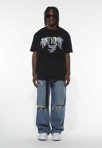 Sort grafisk T-shirt med teksten "LOST YOUTH" og farverige accenter, parret med slidte lyseblå jeans. Fodtøj: hvide sneakers.