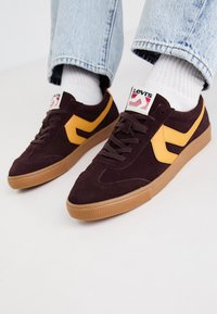 Dunkelburgunderfarbene Wildledersneaker mit gelben Lederakzenten, Gummisohle und weißen Schnürsenkeln. Branding auf der Zunge sichtbar.