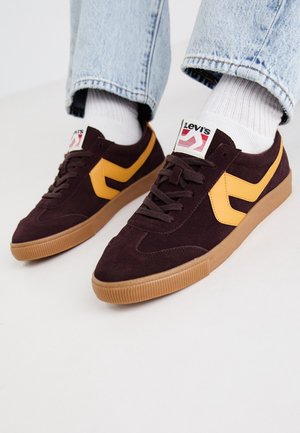 Baskets en suède bourgogne foncé avec des accents en cuir jaune, semelle en gomme, et lacets blancs. Marque visible sur la languette.