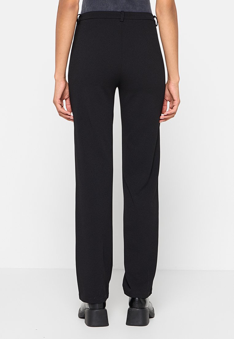 Pantalons noirs à jambes droites, fabriqués dans un tissu lisse avec une taille et des passants de ceinture ; portés avec des chaussures noires.