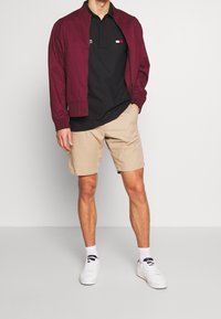 Veste bomber bordeaux sur un polo noir, associée à un short beige et des baskets blanches. Le polo présente un petit logo.