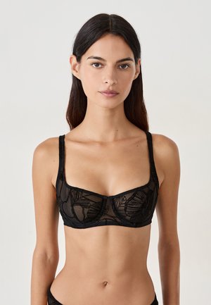 Young woman wearing black sheer mesh bra with floral embroidery, straight dark hair, neutral expression, plain light background.