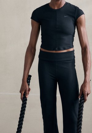 Svart, kortärmad sporttop med hög krage, tillsammans med svarta tajta byxor, som håller i tjocka, tvinnade träningsrep.