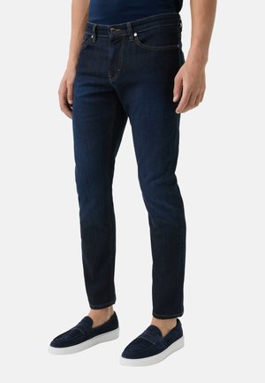 Dark blue denim jeans with a slim fit, featuring yellow stitching and classic five-pocket design. Paired with navy suede slip-on shoes.