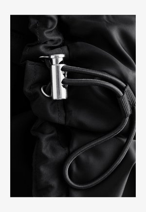 Kovová poistka na šnúrky, ktorá zabezpečuje dve čierne elastické šnúrky na zozberej čiernej látke.