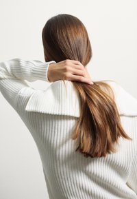 Witte gebreide trui met een brede kraag, met ribbelstructuur. Lang, recht bruin haar is vastgebonden in de nek.