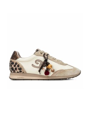 Zapatilla beige y blanca con detalles de estampado de leopardo y cuentas decorativas marrones y doradas sujetas a los cordones sobre un fondo blanco.