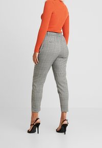 Grå rutiga tapered byxor med elastisk midja, tillsammans med en orange ribbad tröja och svarta högklackade sandaler. Ren bakgrund.