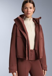 Rövidített, sötétbarna kabát kapucnival, nagy elülső zsebekkel és patentos záródással, amelyhez bordázott krémszínű pulóver társul. Simas, könnyű textúra.