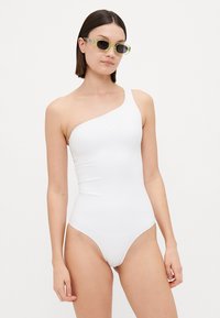 Maillot de bain blanc à une épaule avec une coupe ajustée. Texture lisse, jambes décolletées et détail logo discret à l'avant. Complété par des lunettes de soleil vertes.