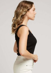 Débardeur noir à côtes avec de larges bretelles, coupe ajustée, porté avec un pantalon taille haute de couleur claire ; texture lisse, vue de côté.