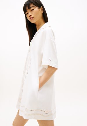 Kvinde iført en løs hvid kjole med korte ærmer og små udskårede blomsterprint, hånden i lommen, kigger mod kameraet.