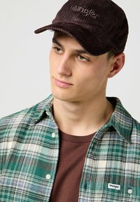 Braune Corduroy-Mütze mit gesticktem Logo, kombiniert mit einem grün-weißen karierten Hemd und einem braunen T-Shirt.
