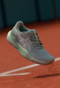 Chaussure de sport gris clair avec une tige perforée, des lacets et une semelle extérieure translucide, suspendue dans les airs au-dessus d'une surface en argile.