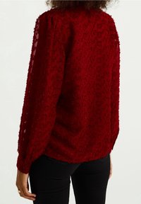Rote, transparente Bluse mit strukturierten Blumendrucken, langen Ärmeln und elastischen Bündchen, getragen über schwarzen Hosen. Weicher, leichter Stoff.