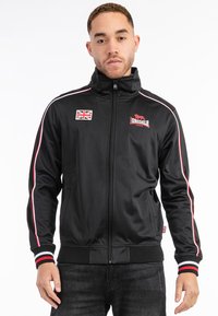 Svart zip-up jacka med randiga röda och vita detaljer, med en brittisk flagga-patch och logotyp. Slät tyg med hög krage och ribbade muddar.
