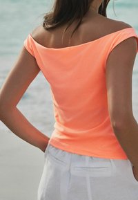 Lyhythihainen, olkapäältä avonainen toppi kirkkaan oranssista kankaasta, yhdistettynä vaaleisiin valkoisiin housuihin. Sileä pinta ja vartalonmyötäinen muotoilu.