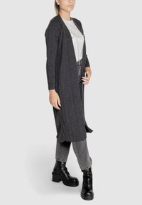 Cardigan lungo aperto grigio scuro in tessuto a coste, con maniche lunghe. Indossato sopra una t-shirt bianca e abbinato a stivali neri alla caviglia.