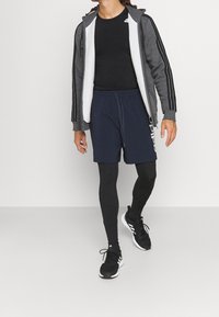 Mann, der in schwarzen Sportleggings, blauen Shorts, schwarzem T-Shirt, grauem Kapuzenpullover und schwarzen Sneakers mit weißen Sohlen geht.
