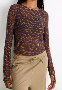 Långärmad topp i mörkblått med ett orange abstrakt mönster, med en figurnära design och tumhål vid ärmsluten. Kombinerad med beigefärgade ribbade byxor.