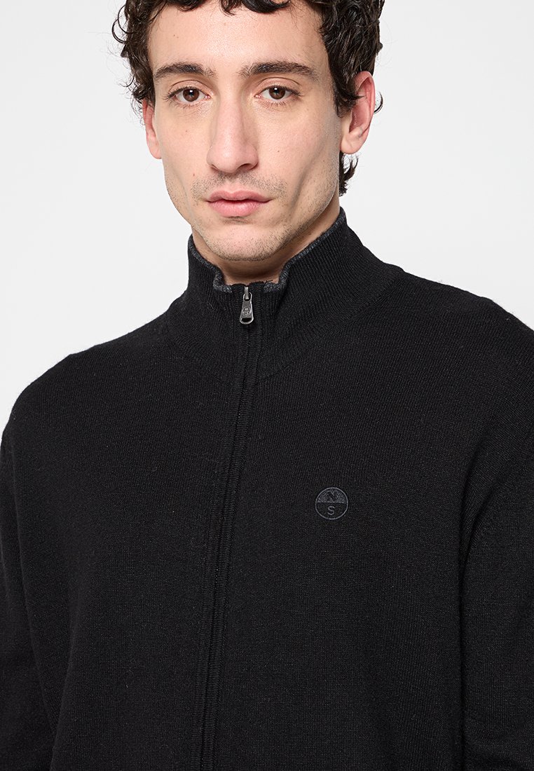 Joven con cabello corto y rizado, vistiendo un suéter negro con cremallera y un pequeño logotipo bordado en el pecho, contra un fondo liso.