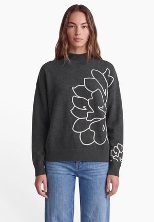 Kvinde med langt brunt hår iført en mørkegrå sweater med stort hvidt blomsteromrids og blå jeans, stående mod en hvid baggrund.