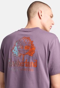 La camiseta de algodón morada presenta un gran gráfico del logo multicolor de Timberland en la parte posterior, con un diseño de árbol y el texto "Rugged Active Gear."