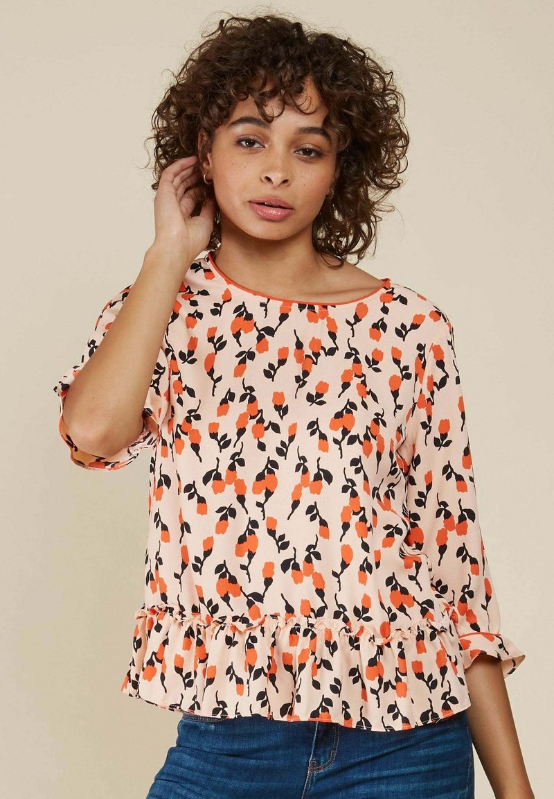 Haut imprimé floral avec un fond pêche, présentant des fleurs noires et orange, un ourlet volanté, et des manches trois-quarts avec un col en scoop.