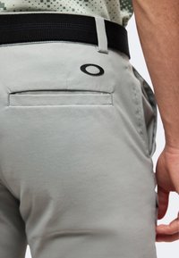 Κοντινό πλάνο ενός ατόμου που φοράει ανοιχτό γκρι παντελόνια με πίσω τσέπη και μαύρη ζώνη, λογότυπο κοντά στην τσέπη και μέρος του βραχίονά του ορατό.