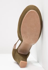 Chaussure à talon en daim marron montrant la semelle en caoutchouc à motif floral et le petit talon carré sur une surface blanche.