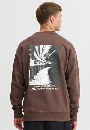 Bruine sweatshirt met een gestructureerde afwerking heeft een zwart-wit fotografische print op de achterkant, gelabeld "©1999 SOLID ARCHIVE ALL RIGHTS RESERVED."