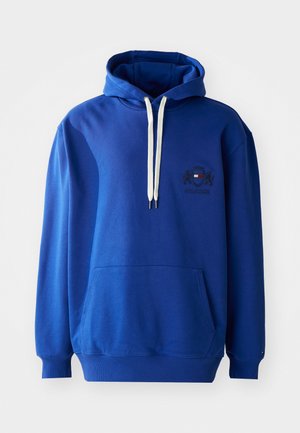 Sininen huppari, joka on valmistettu pehmeästä kankaasta. Siinä on etutasku, kontrastiväriset valkoiset nyörit ja pieni brodeerattu logo rinnassa.